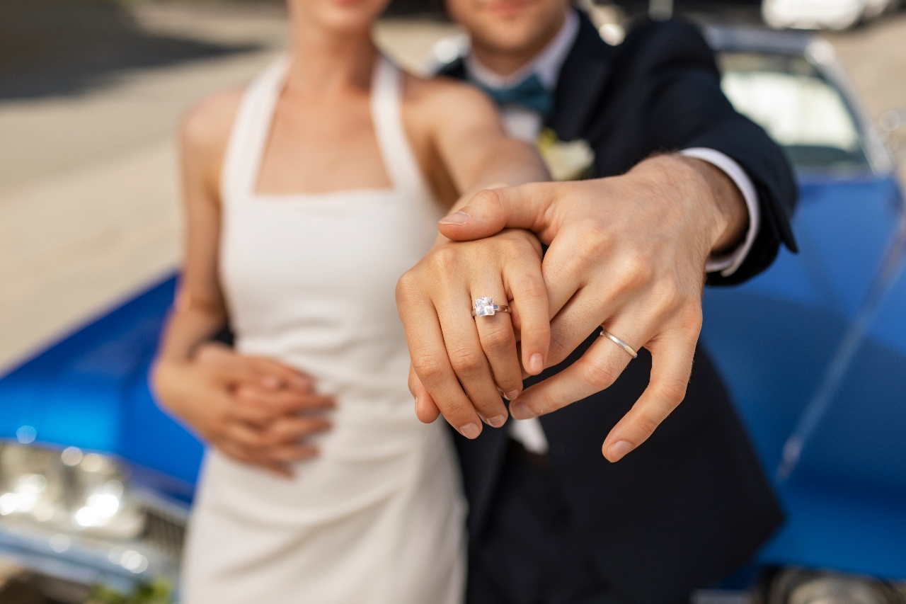 A couple poses leaning against a blue vintage car, showcasing their wedding rings.