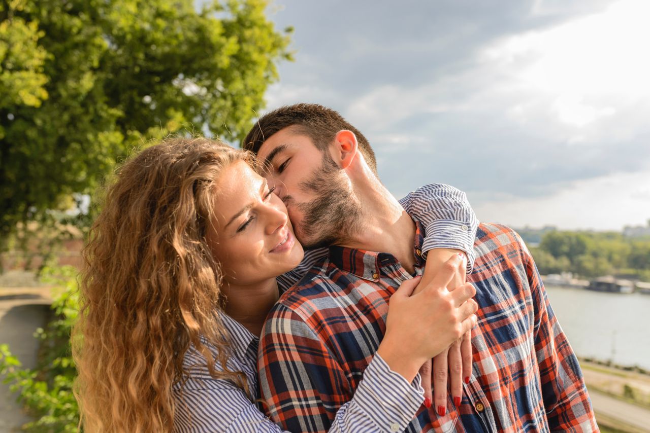 A woman embraces a man from behind in a park as he kisses her on the cheek.
