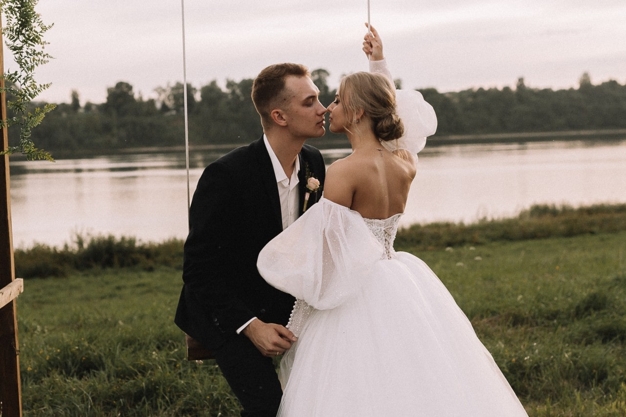 A bride and groom on their wedding day, the groom seated on a swing while the bride leans in for a kiss, with a large body of water in the background.