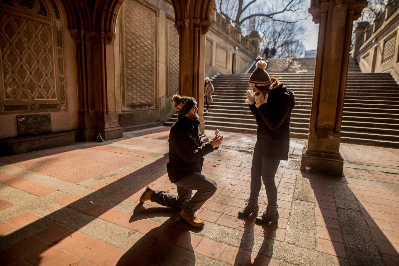 Man kneeling and proposing to woman in winter clothes under an ornate archway.