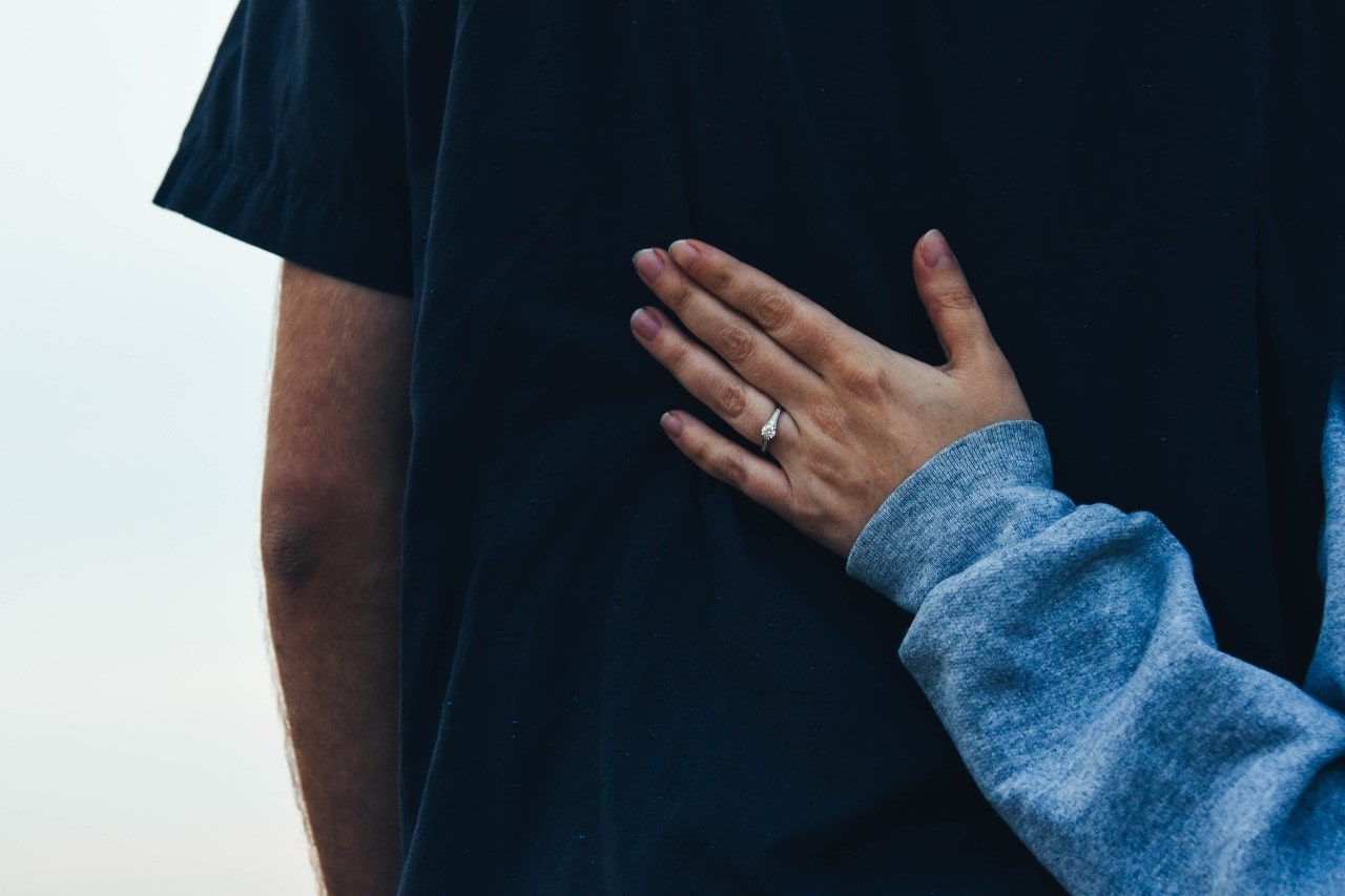 A close-up of a woman’s hand resting on a man’s back, showcasing her beautiful engagement ring.