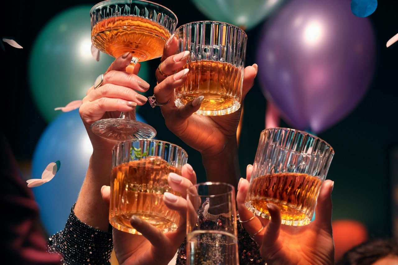 A close-up of people’s hands as they raise their glasses, surrounded by balloons and confetti.