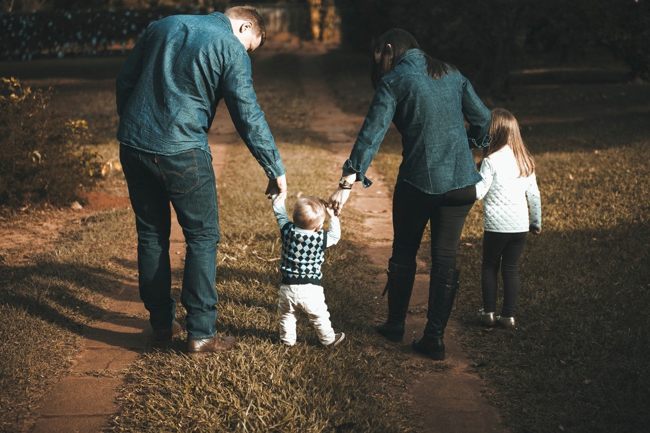 A family of four walks down a grassy path, with the parents holding their toddler’s hands while their older child walks beside them.