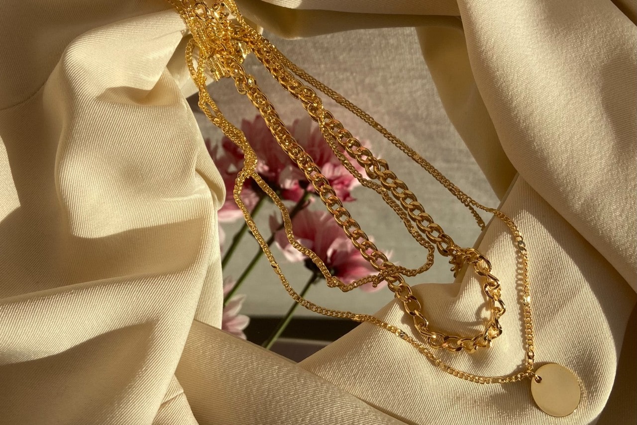 A set of layered gold necklaces displayed on cream fabric with a blurred pink flower in the background.