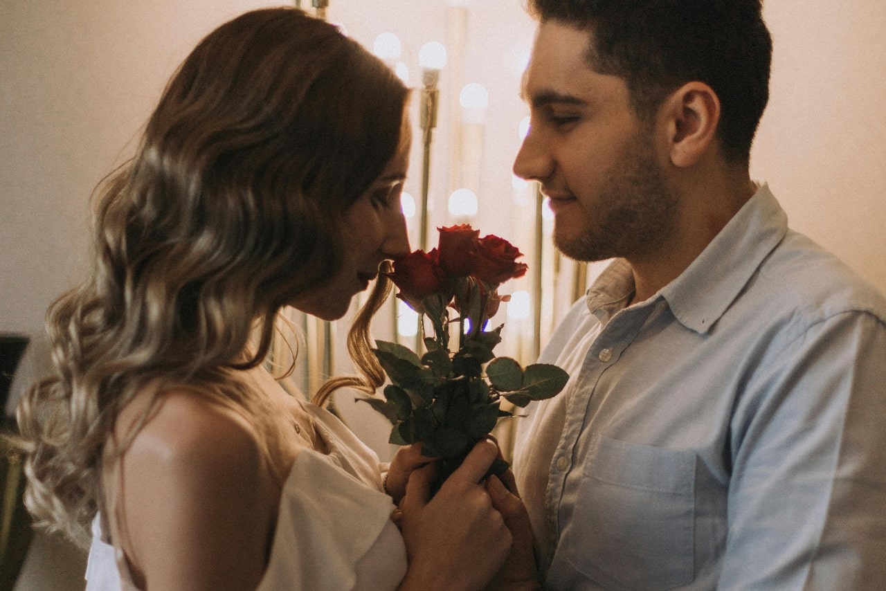 A man and woman standing in front of each other, while the woman smells a small bouquet of red roses.