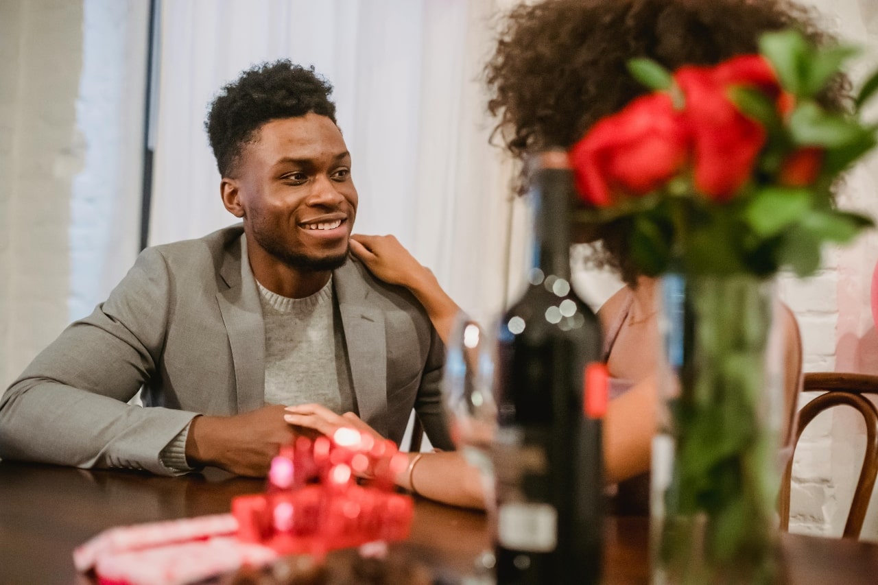 A man and woman sitting at a fancy table with a bottle of wine and a vase with red roses nearby.