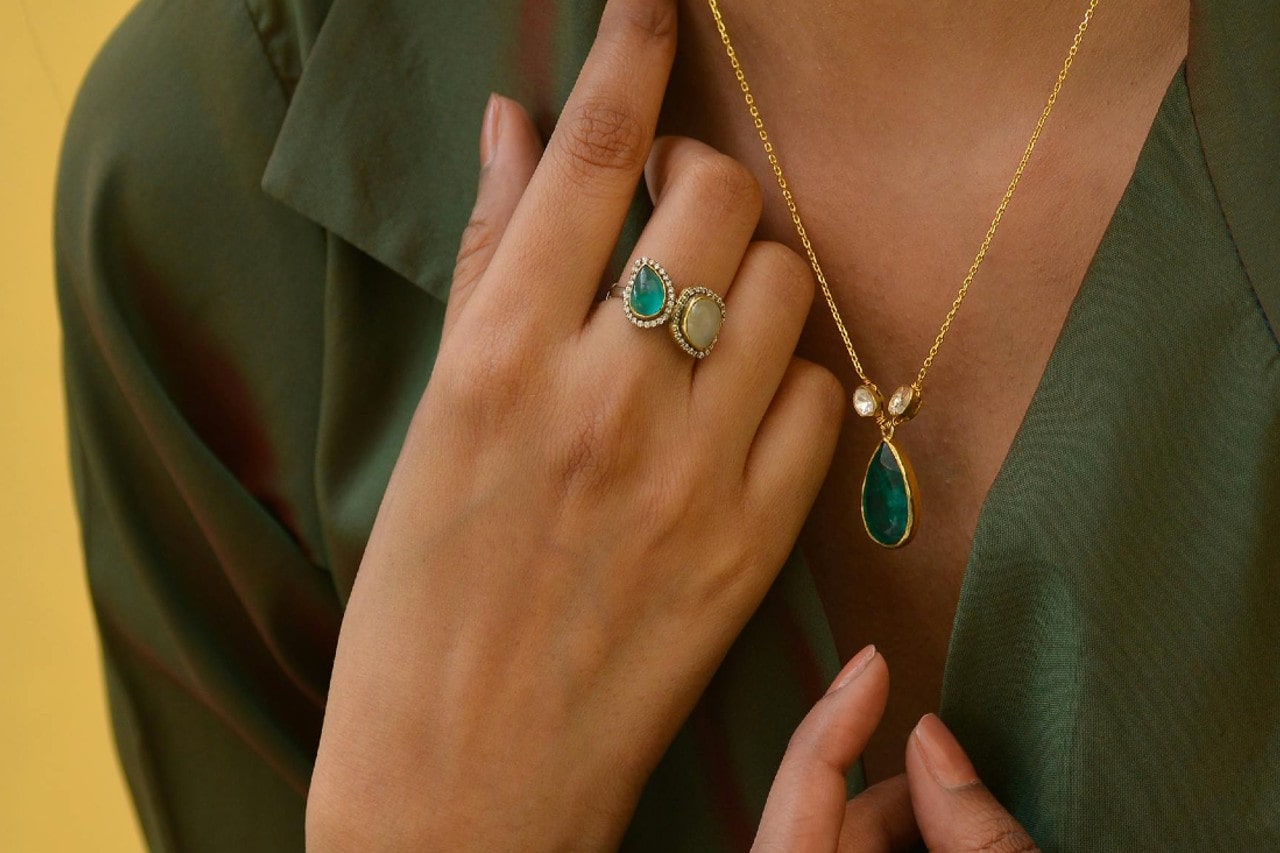 A woman wearing a green shirt rests her hand on her chest, showcasing a gold necklace with a green teardrop pendant and a ring with a similar green stone.