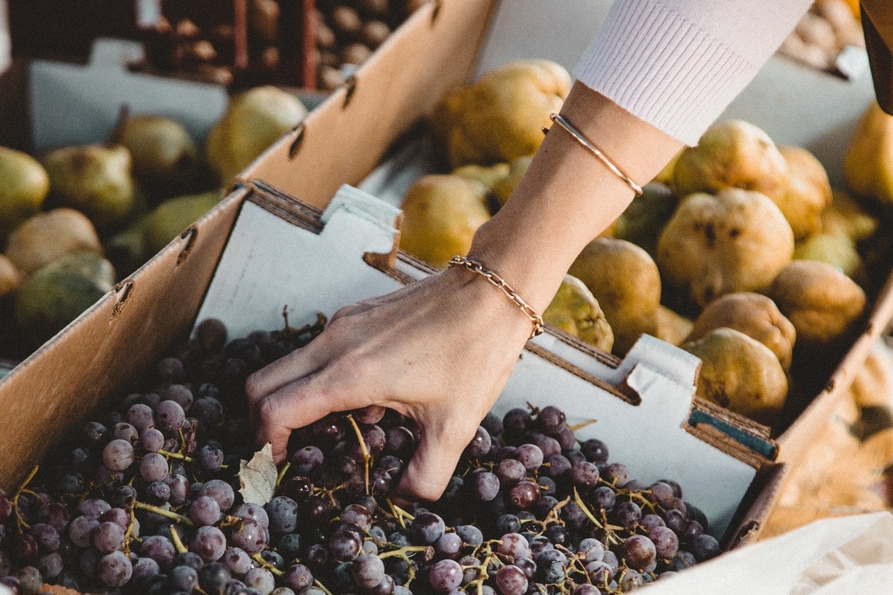 A woman's hand adorned with delicate gold bracelets as she picks fresh grapes from a market stall filled with fruit