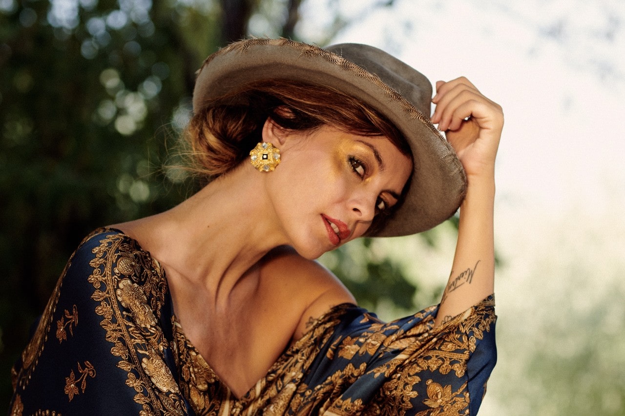 A woman with a hat tipping over her face, wearing golden earrings and blue patterned dress, and looking away from the camera.