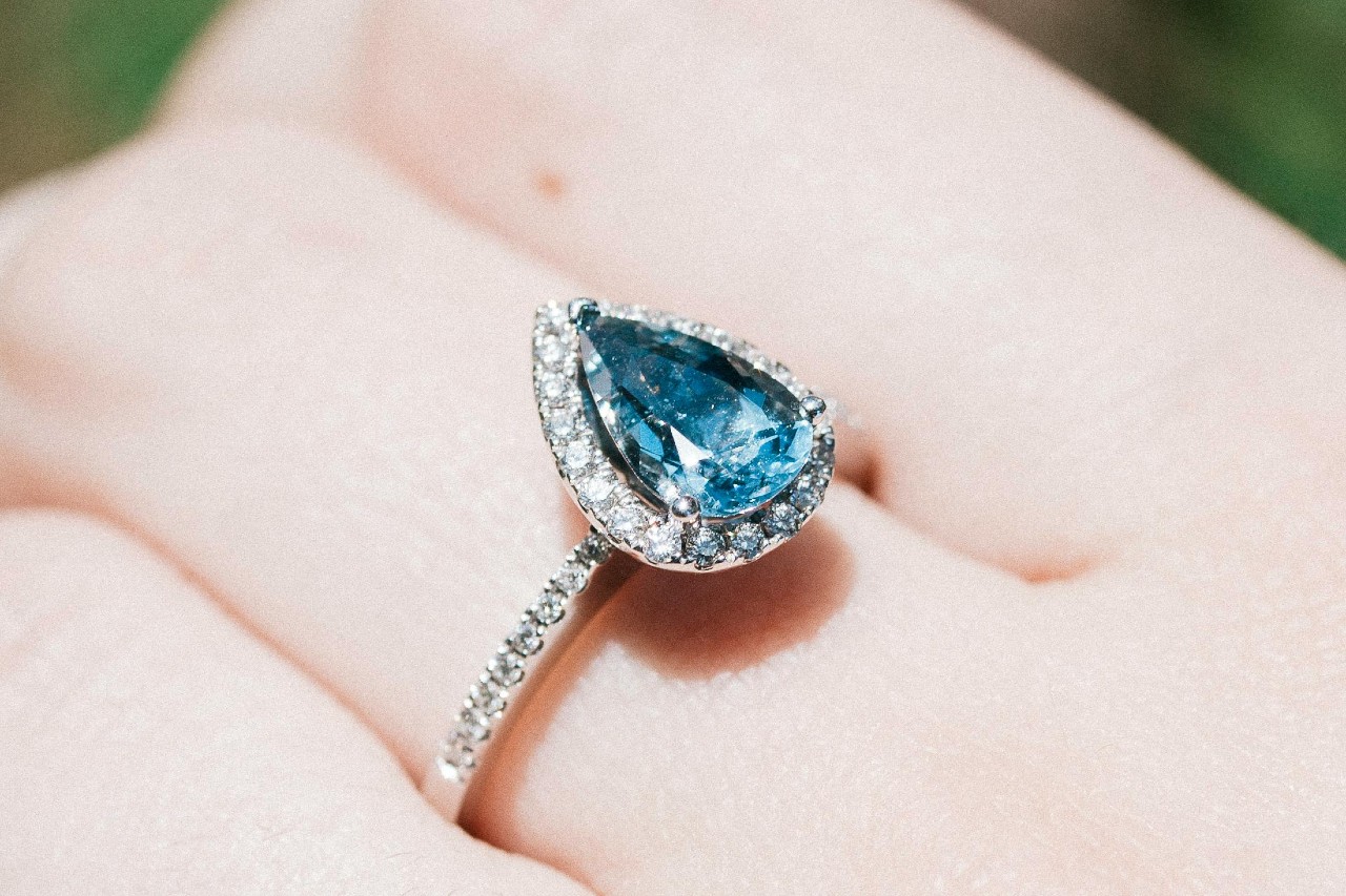 A close-up of an elegant halo ring featuring a pear cut Yogo sapphire center stone on a woman’s finger.