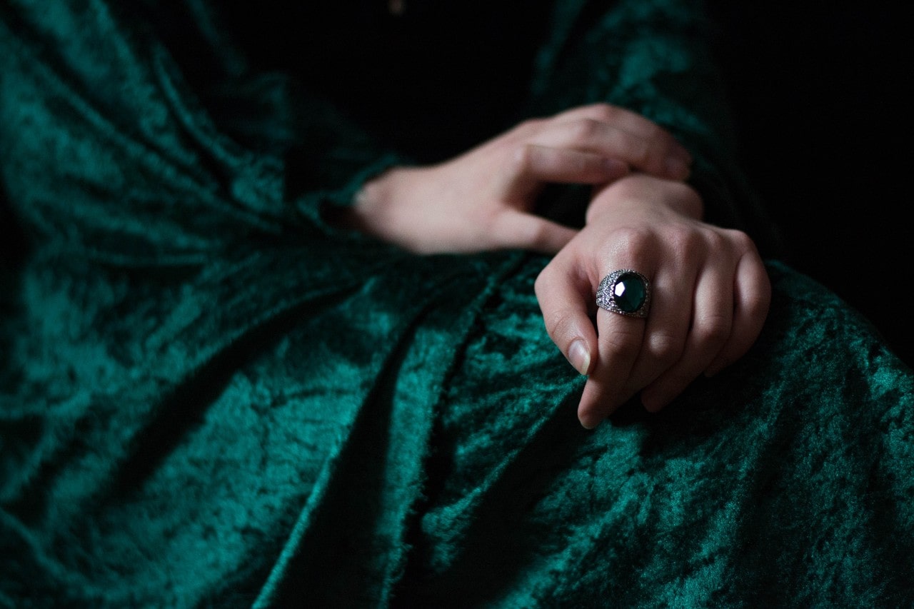 A woman in a deep green velvet dress showcasing an ornate statement fashion ring with a large oval emerald.