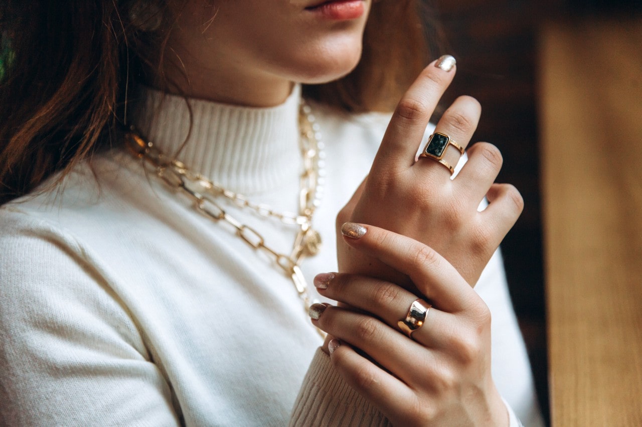 A close-up of a woman’s hands and neck adorned with chunky gold rings and layered chain necklaces over a white turtleneck.