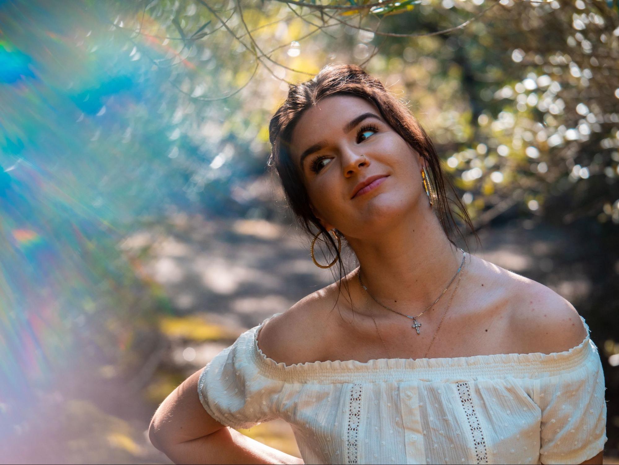 A woman in a white dress wears hoop earrings, a layered diamond cross necklace, and a nose piercing, against a soft, blurred floral background. A woman in a white dress wears hoop earrings, a layered diamond cross necklace, and a nose piercing, against a soft, blurred floral background.