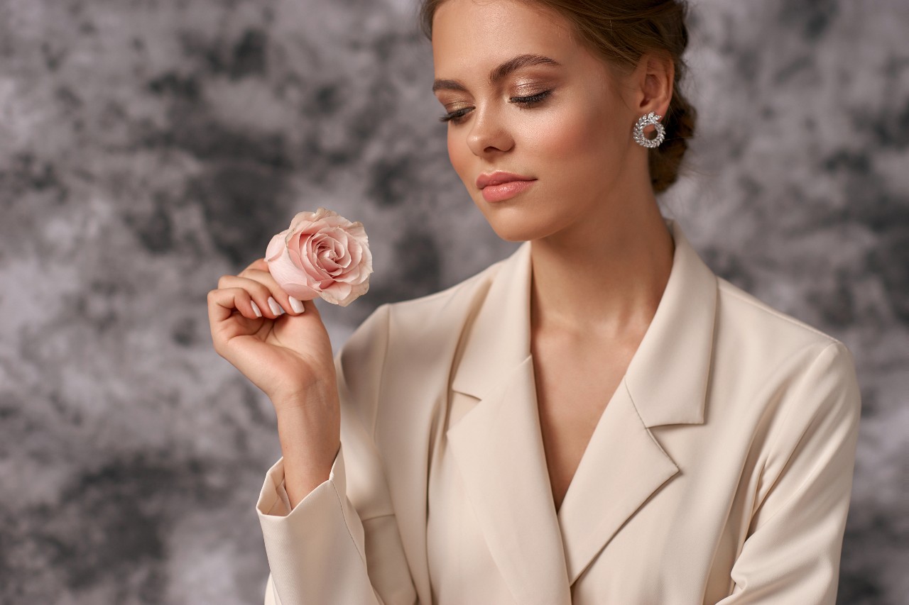 A woman in a beige blazer wearing elegant hoops, holds a pink rose, against a soft gray background.