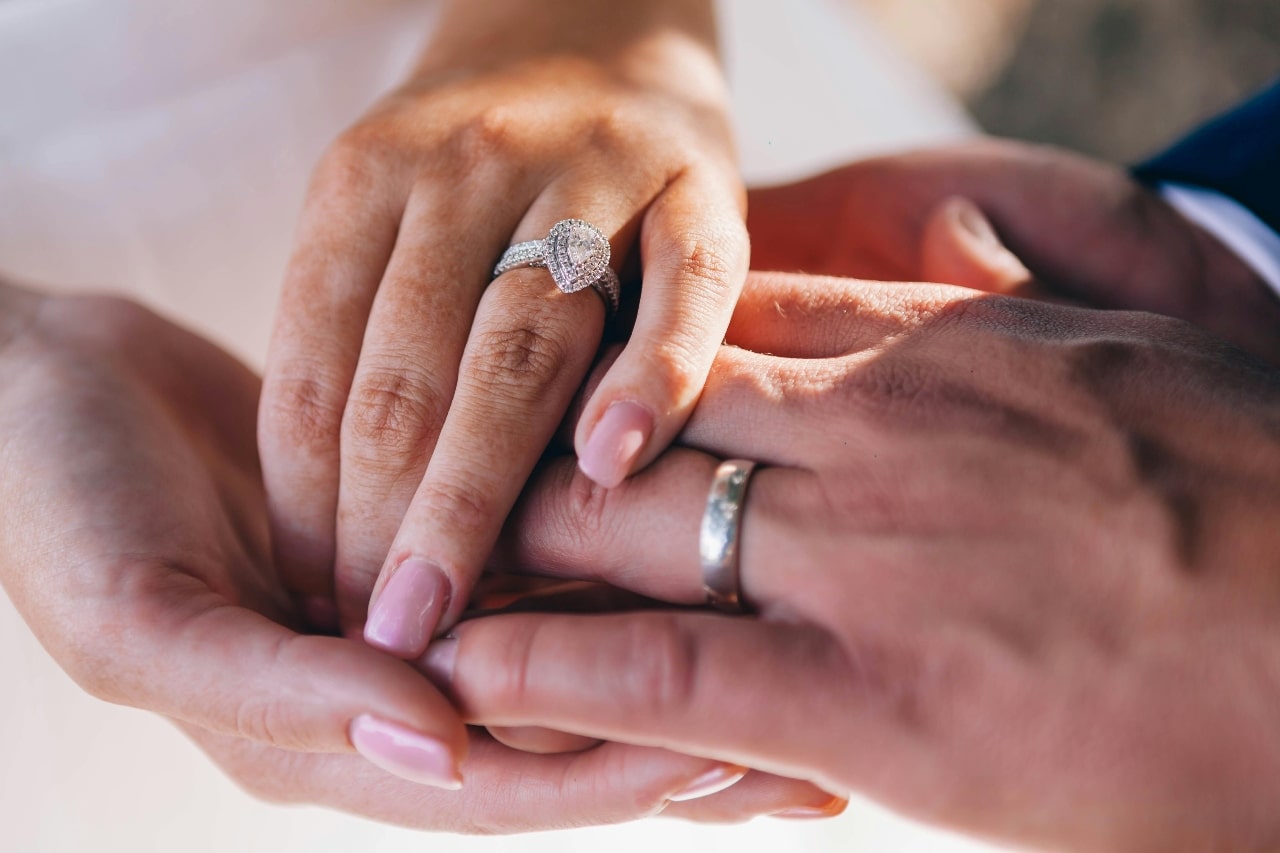 A close up of two hands, showcasing a side-stone diamond engagement ring on a woman’s hand.