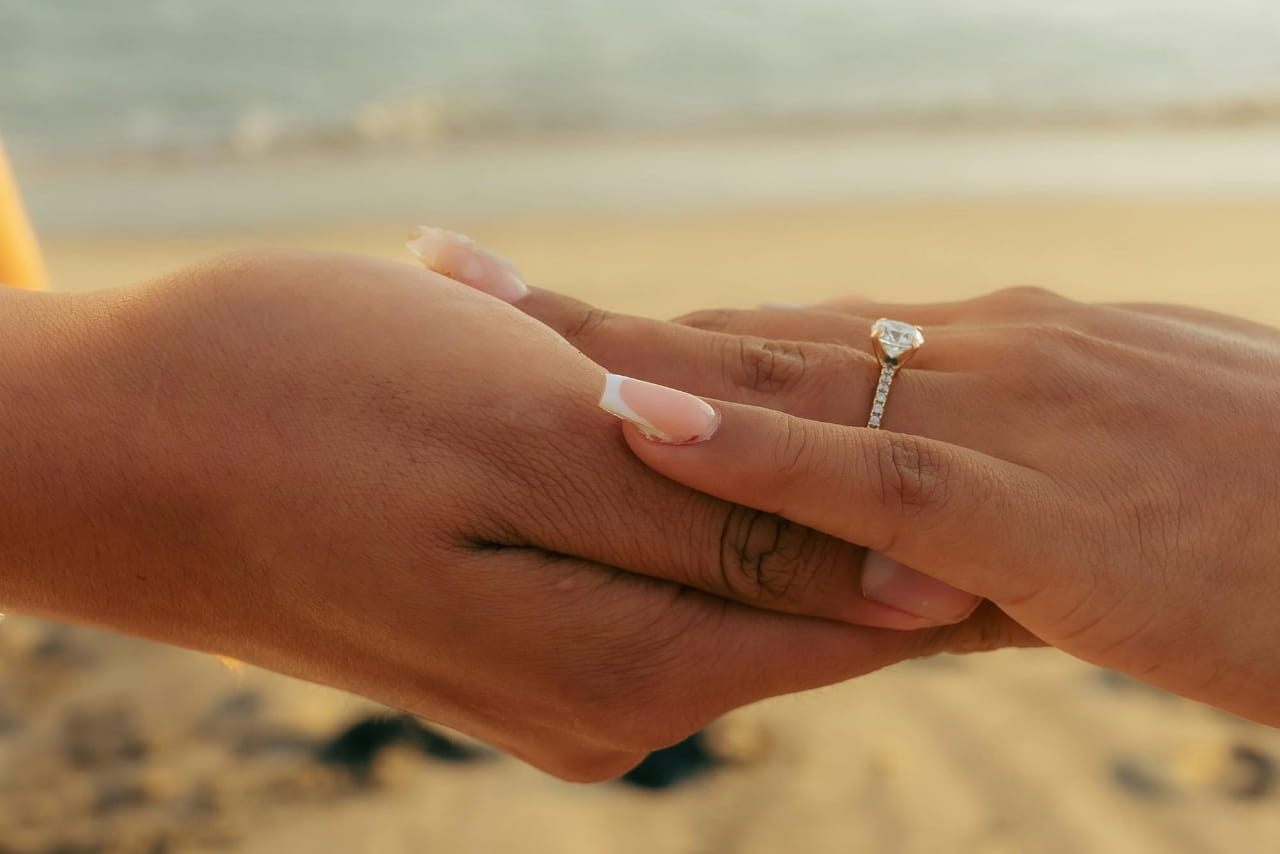 Two hands together in front of the beach, showcasing a side-stone diamond engagement ring on a woman’s finger with a french manicure.