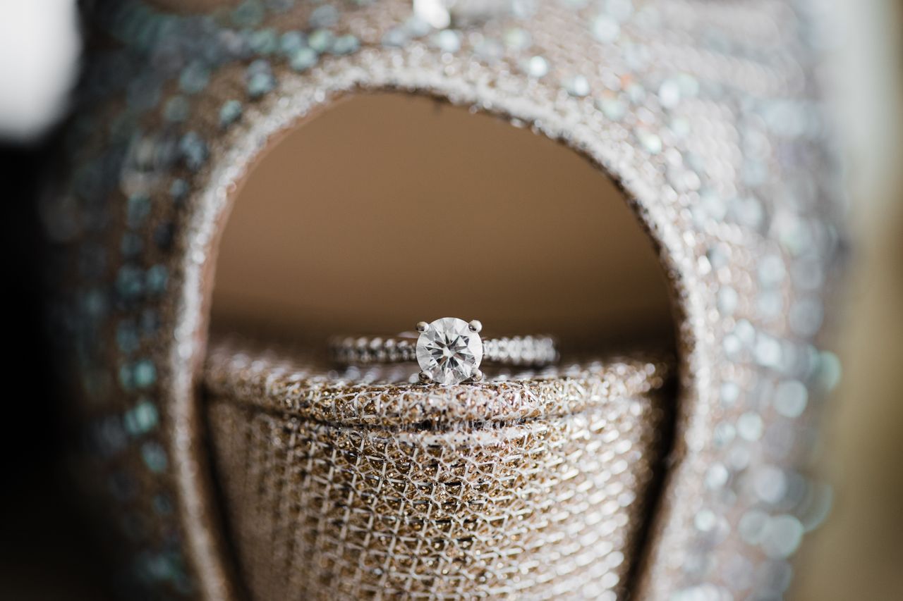A close-up of a white gold round cut side stone engagement ring placed on the edge of an open-hill shimmering bridal shoe.