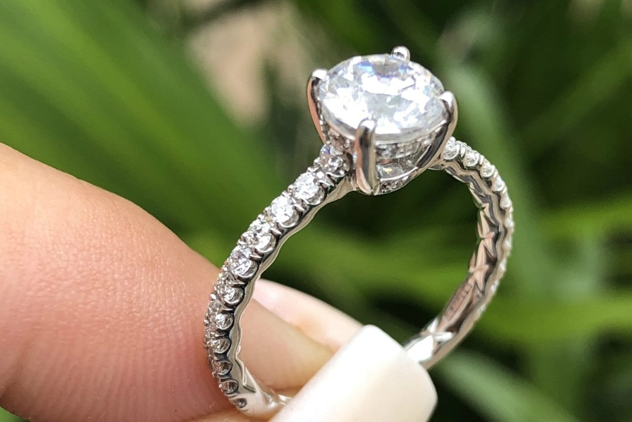 A close-up of a white gold four prong diamond sidestone engagement ring with cushion cut center diamonds against a blurry grass background.
