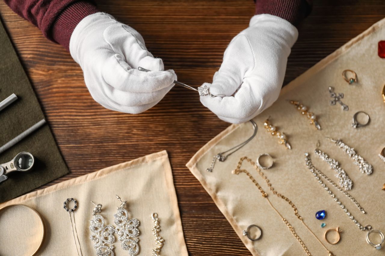 A jeweler wearing white gloves inspects a diamond ring over a wooden table, with various pieces of elegant jewelry displayed on beige cloth. A jeweler wearing white gloves inspects a diamond ring over a wooden table, with various pieces of elegant jewelry displayed on beige cloth.