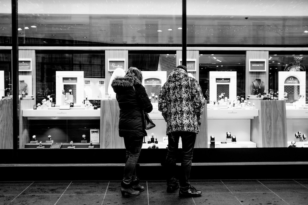 Two people in winter coats stand close together, admiring a brightly lit jewelry display through a large window in a black-and-white setting. Two people in winter coats stand close together, admiring a brightly lit jewelry display through a large window in a black-and-white setting.