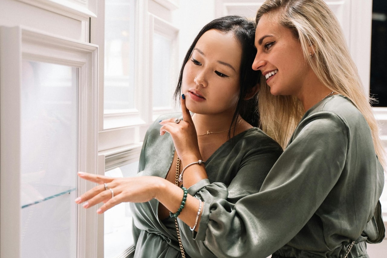 Two women in matching green outfits examine jewelry in a display case at a luxurious boutique, one pointing while the other smiles. Two women in matching green outfits examine jewelry in a display case at a luxurious boutique, one pointing while the other smiles.