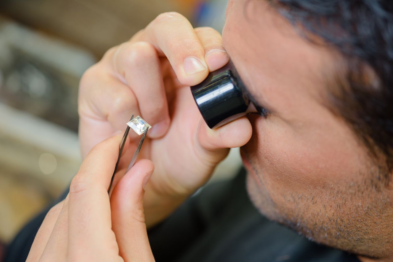 A jeweler looking through a small magnifying glass at a princess cut diamond.