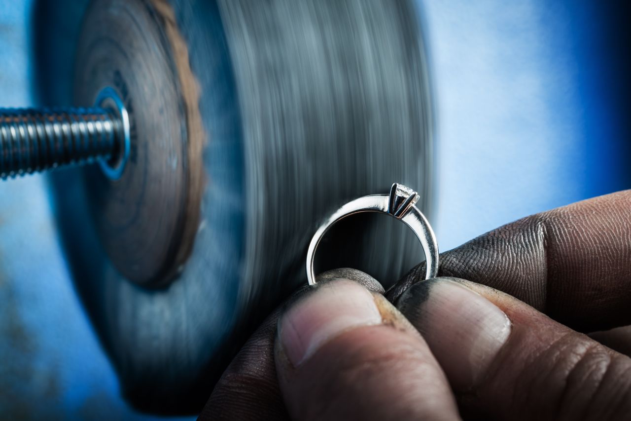 A close-up of an engagement ring getting sanded, held by a jeweler’s hands covered in residue.