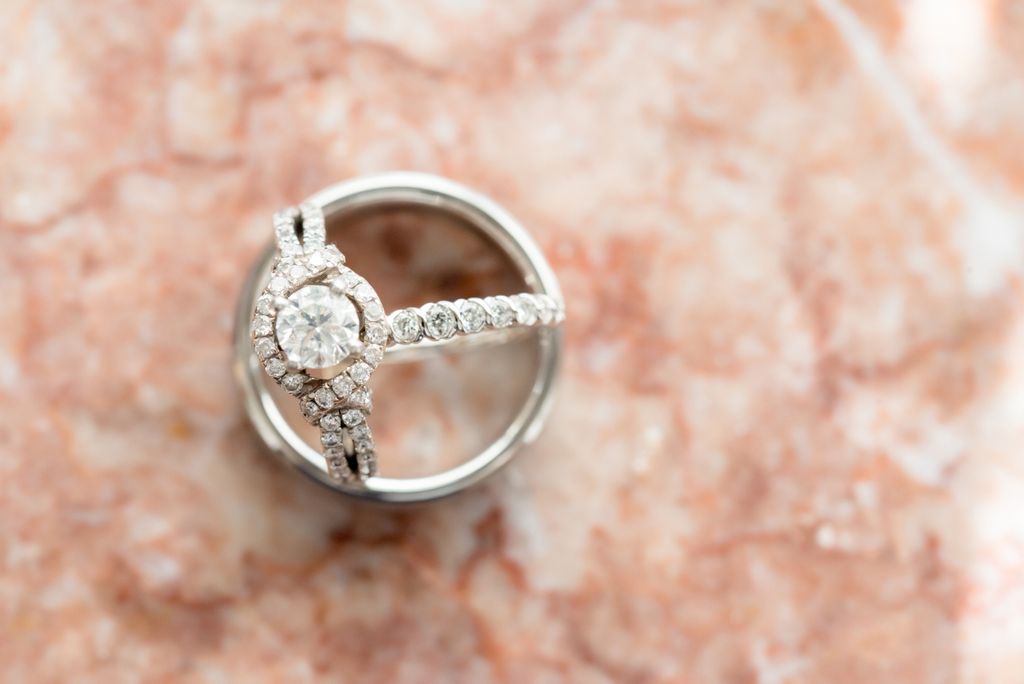 A close-up of a diamond engagement ring on top of a polished yellow gold wedding band on a marble blurred surface.