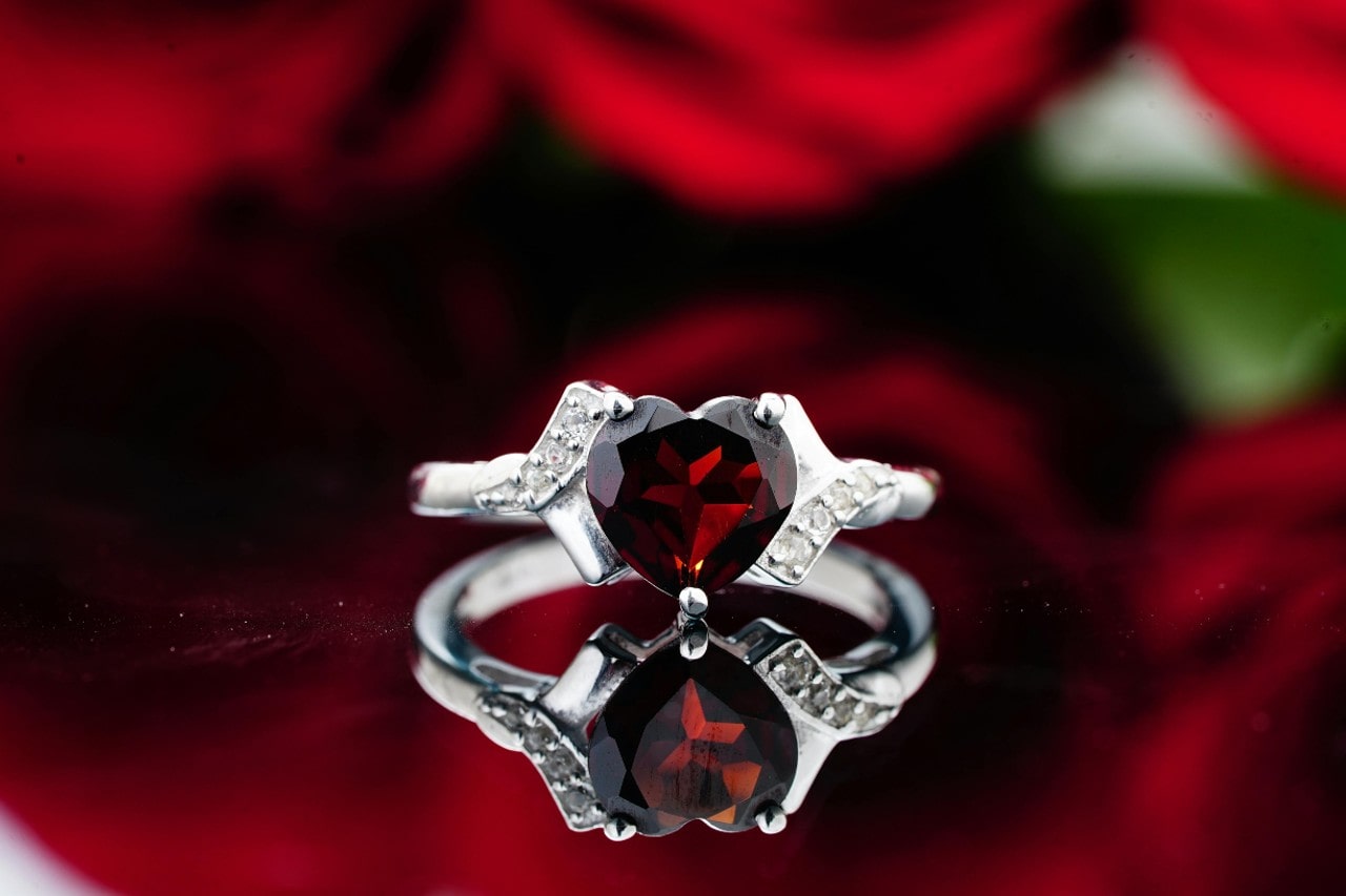 A white gold engagement ring with a heart-shaped ruby flanked by small diamonds resting on a reflective surface, with blurred red roses in the background.