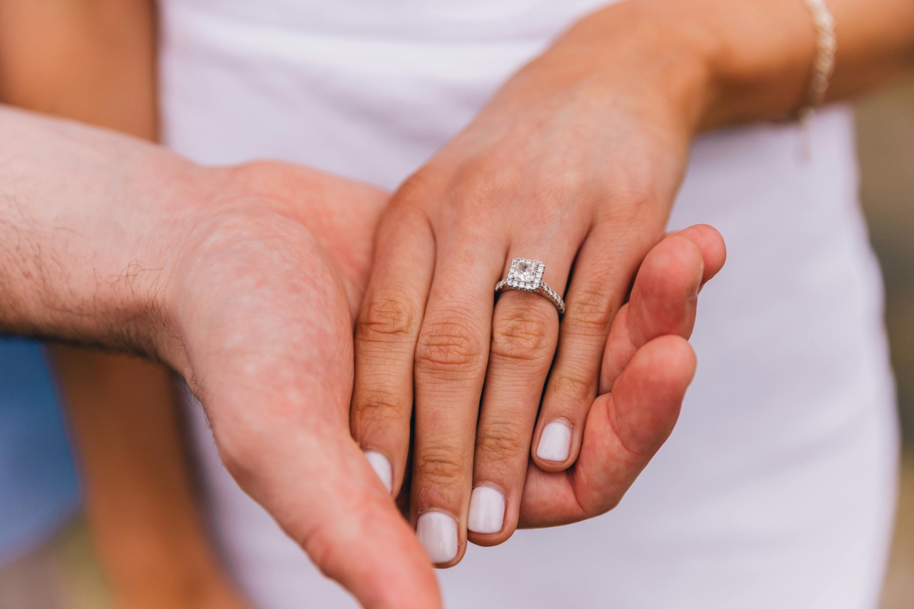 A close-up of a couple's hands, with a bride wearing a sparkling diamond engagement ring featuring a square-cut center stone.