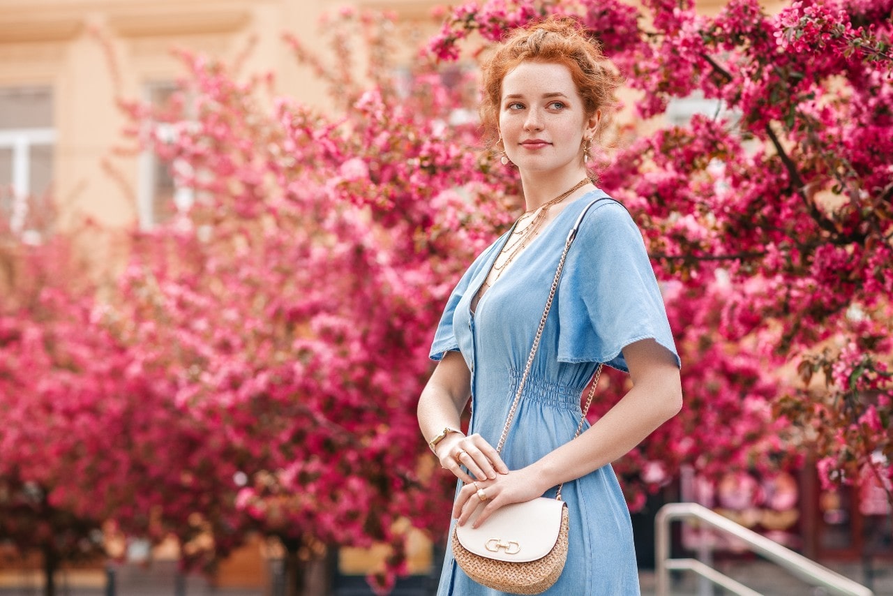 A young woman in blue dress and layered necklaces stands against blooming sakura