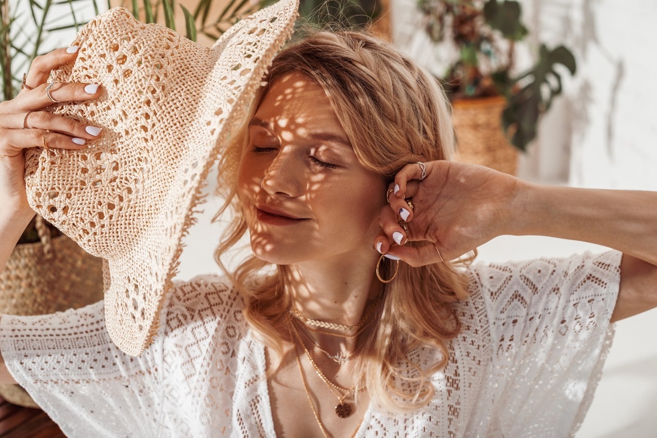 A woman in a white lace outfit in sunlight with her eyes closed, showcasing yellow gold hoops and layered necklaces, and holding a woven straw.