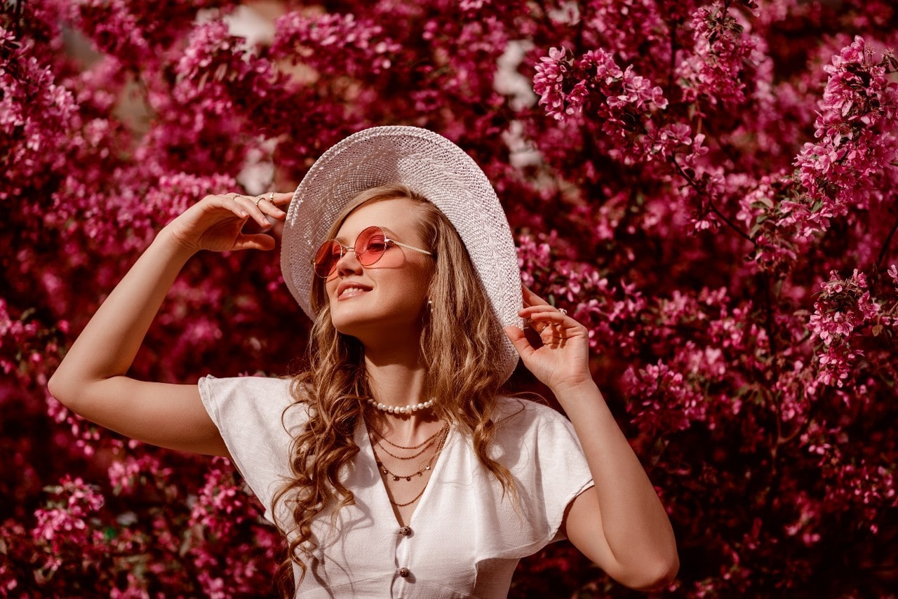 A woman in the white blouse and hat stands against a pink tree, showcasing a layered yellow gold necklace.